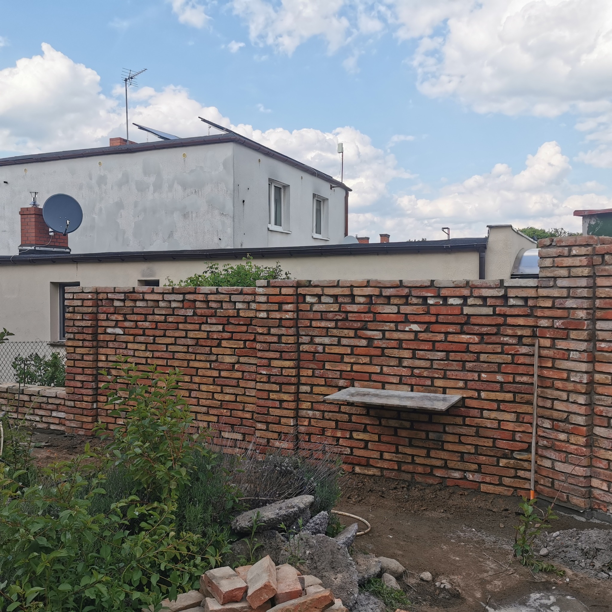 Ceglany murek ogrodowy w trakcie budowy, z widocznym fragmentem budynku w tle i roślinnością w przedniej części kadru. Widoczna zaprawa i niedokończone fragmenty.