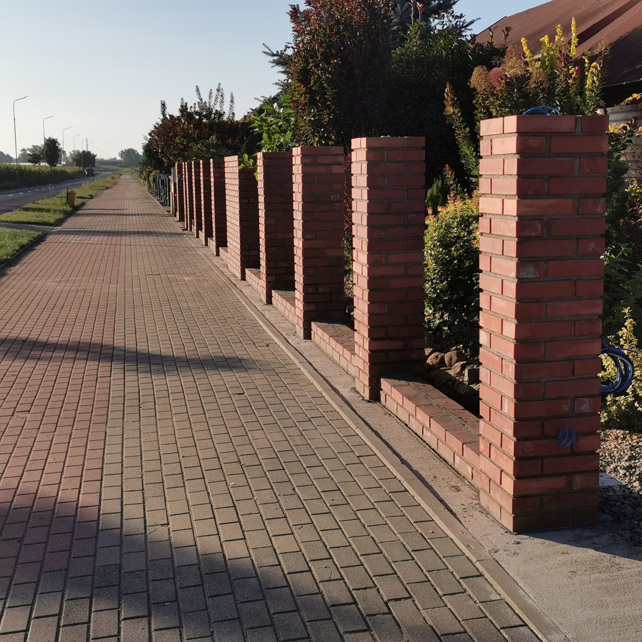 Czerwone, ceglane słupki ogrodzeniowe wzdłuż chodnika z kostki, z zielenią w tle. Widok na perspektywę ogrodzenia i ulicę w oddali w słoneczny dzień.