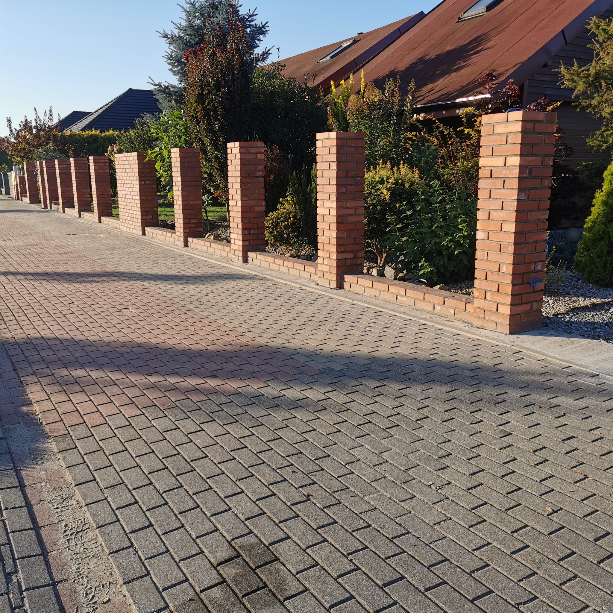 Murowane słupki ogrodzeniowe z cegły klinkierowej, oddzielające posesję od chodnika z kostki brukowej. W tle zieleń i zabudowa jednorodzinna.