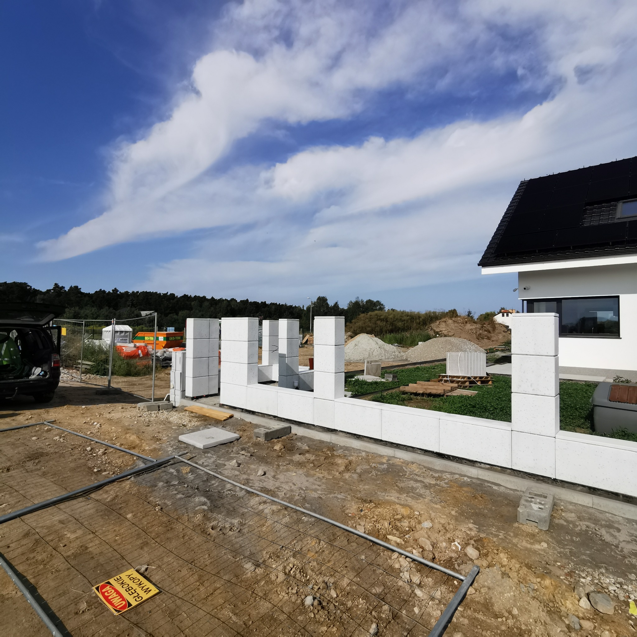 Wznoszenie muru z białych bloczków betonowych, widoczne zbrojenie w gruncie i fragment domu z czarnym dachem na tle błękitnego nieba z chmurami.