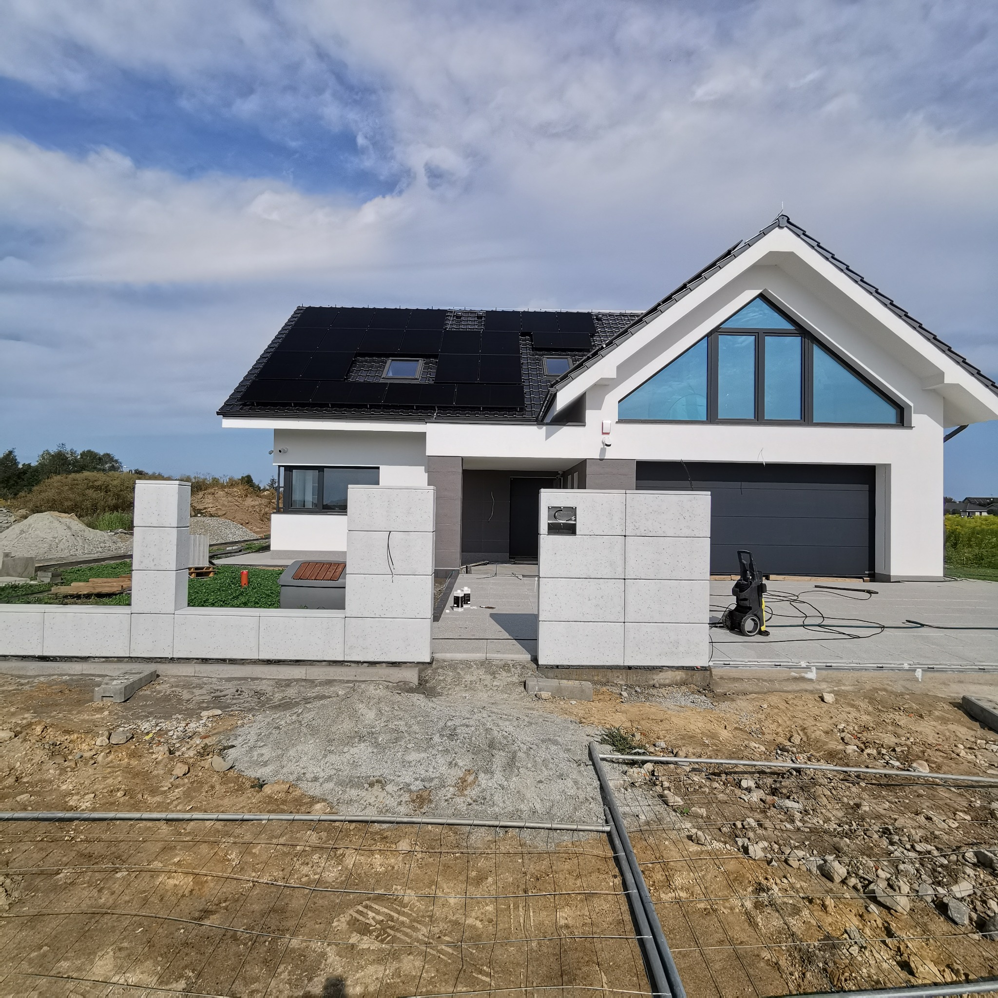 Nowoczesny dom jednorodzinny z elementami ogrodzenia z betonu architektonicznego, widoczny teren budowy i przygotowania do dalszych prac.