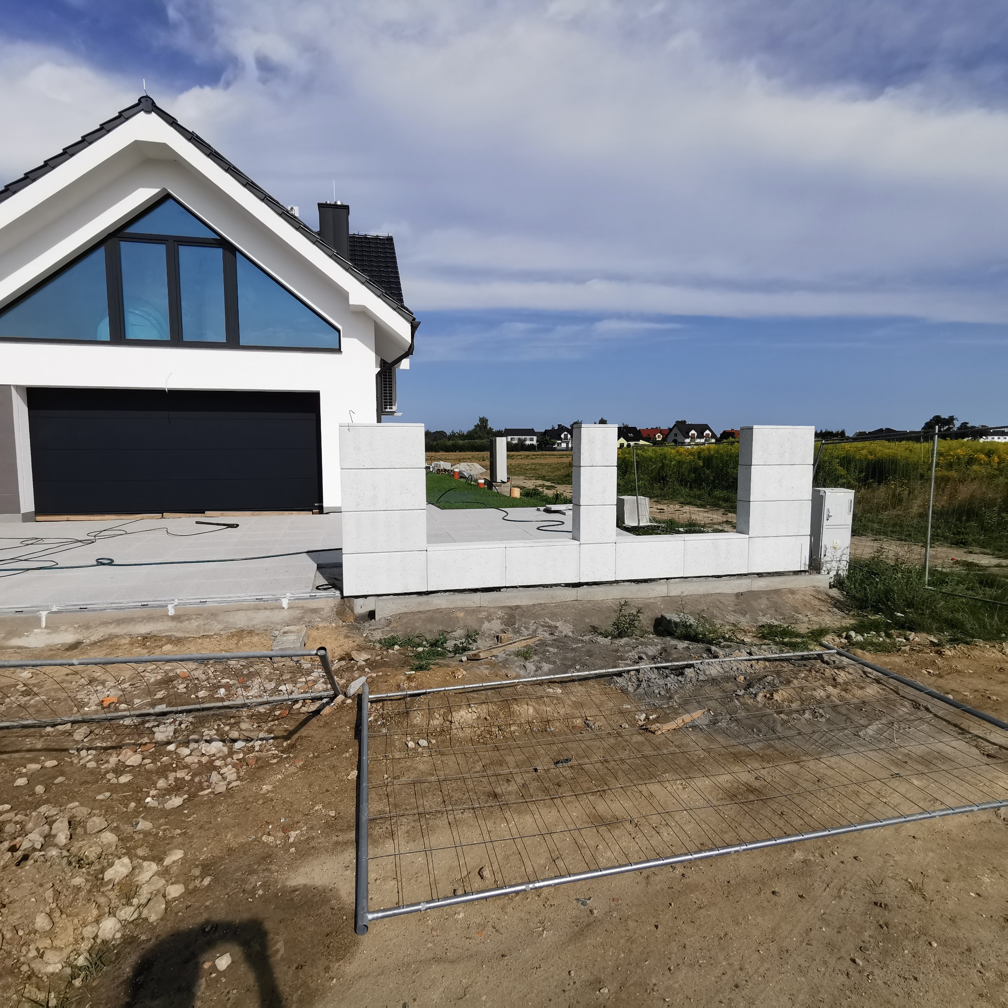 Wznoszenie ogrodzenia betonowego modułowego w Śremie. Widoczne bloczki fundamentowe z betonu architektonicznego i tymczasowe ogrodzenie budowlane.