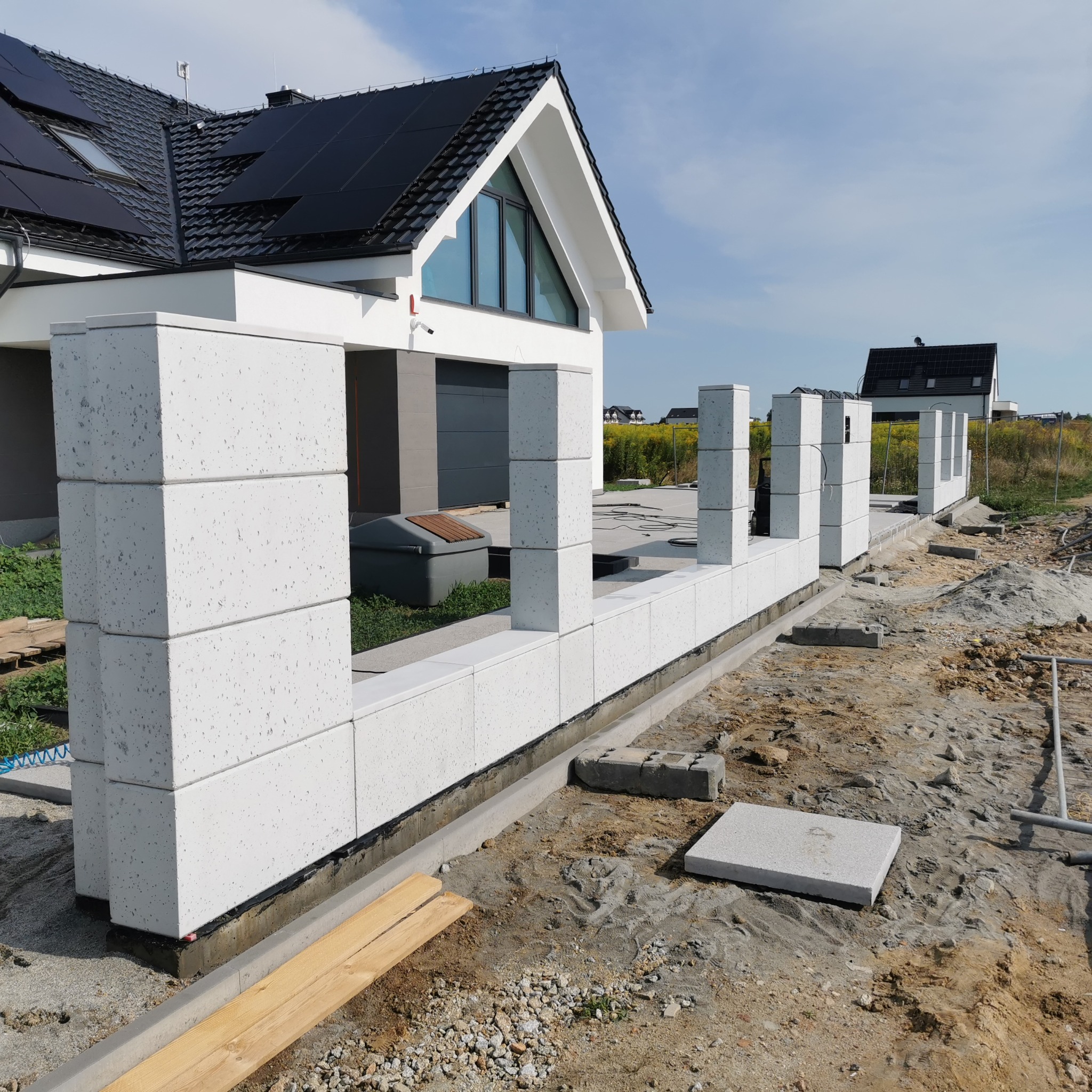Budowa ogrodzenia modułowego z białych bloczków betonowych na posesji z nowoczesnym domem. Widoczny fundament i słupki ogrodzeniowe. Prace w toku.