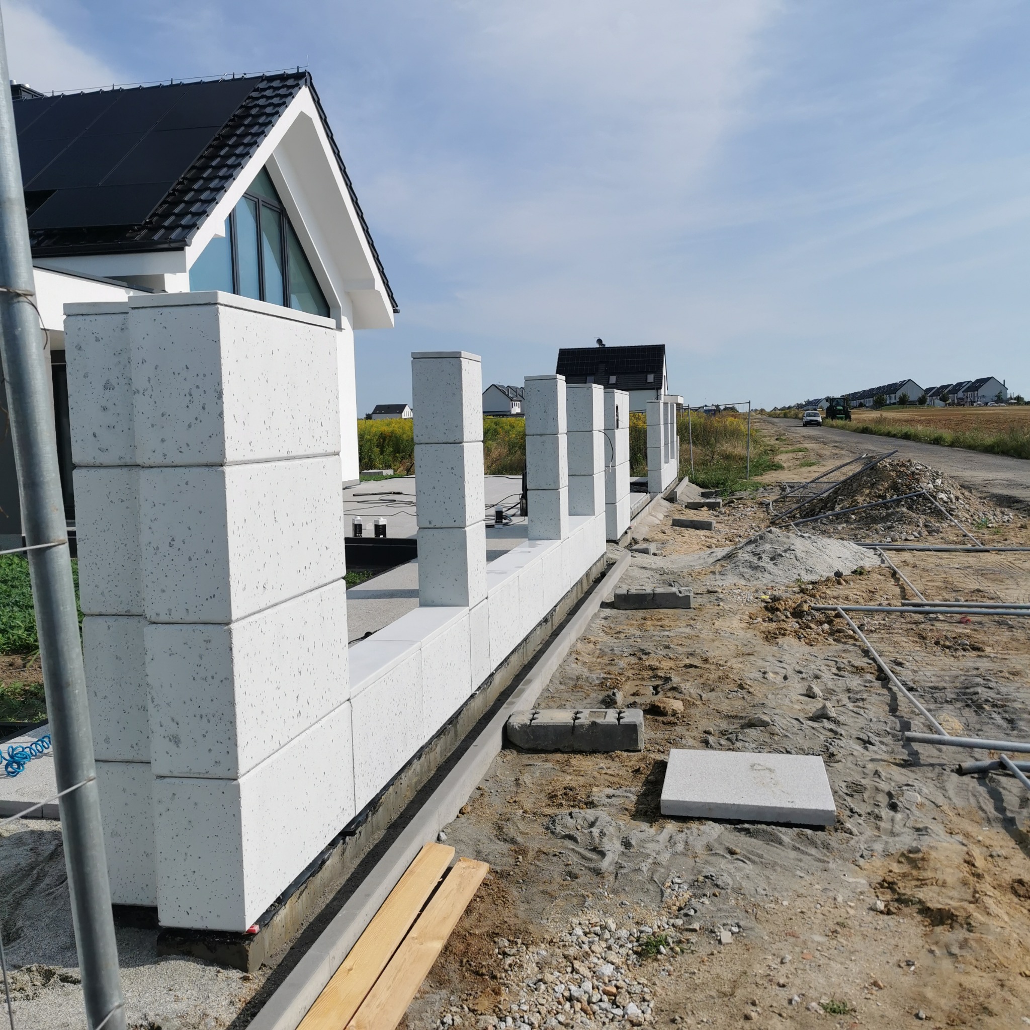 Wznoszenie muru z białych bloczków betonowych, widoczne fundamenty i elementy konstrukcyjne. Budowa ogrodzenia na nowym osiedlu domów jednorodzinnych w słoneczny dzień.