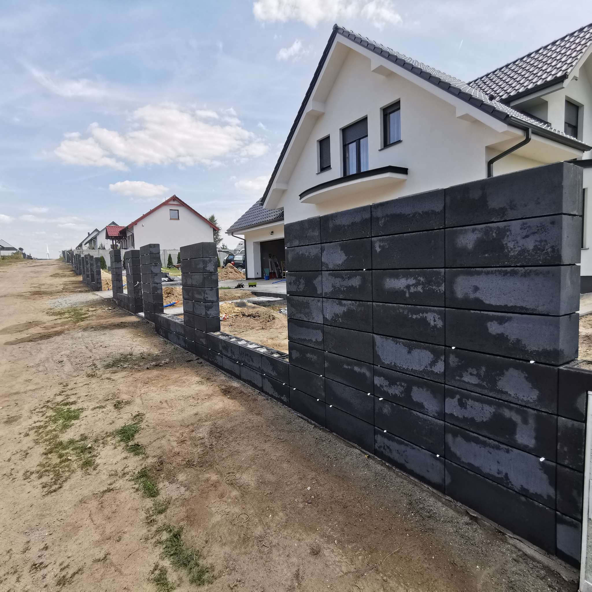 Częściowo ukończony, nowoczesny, grafitowy płot z betonowych bloczków na tle jasnego domu z ciemną dachówką i błękitnego nieba z chmurami. Widoczne kolejne etapy budowy.