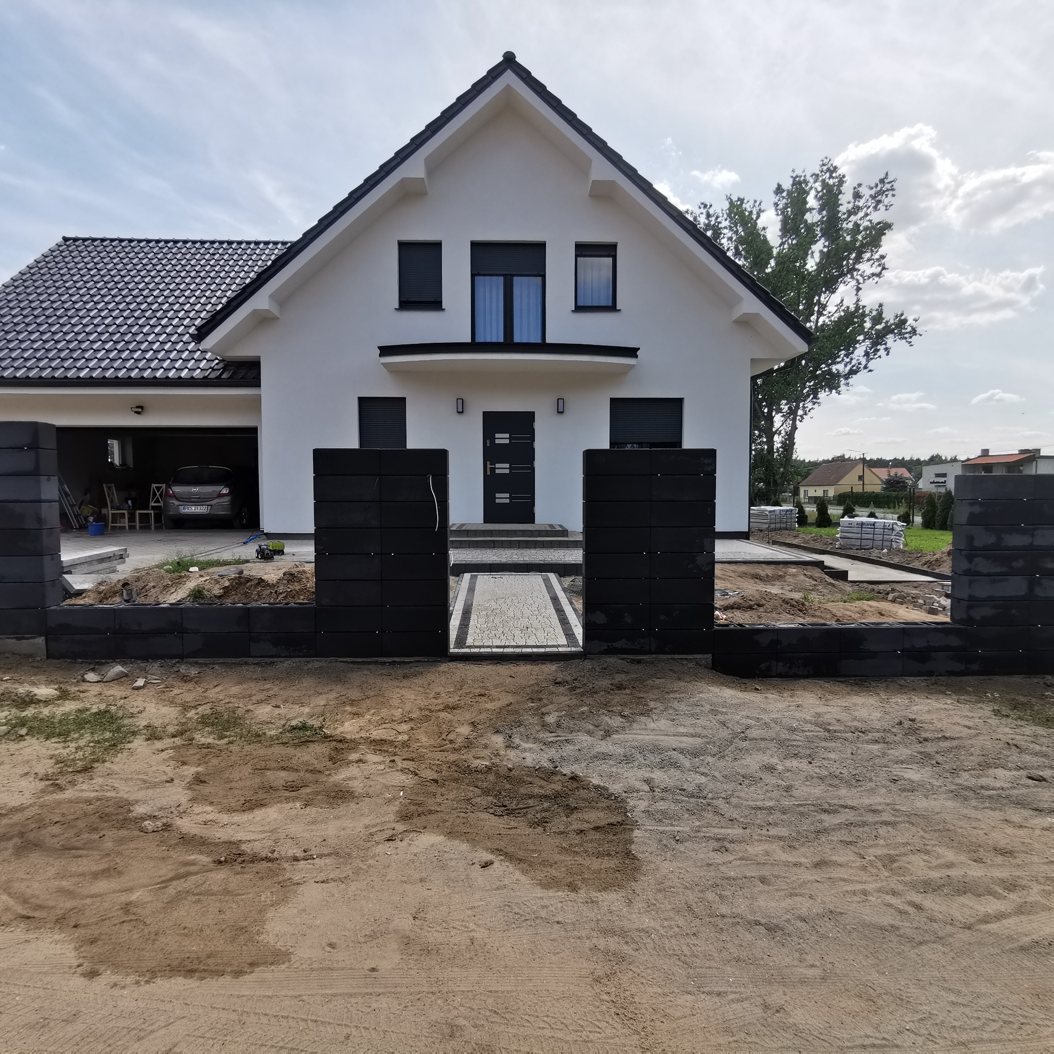 Nowoczesny dom jednorodzinny z czarnym, modułowym ogrodzeniem z pustaków betonowych i ścieżką z kostki. Widok z zewnątrz na posesję w trakcie prac wykończeniowych.