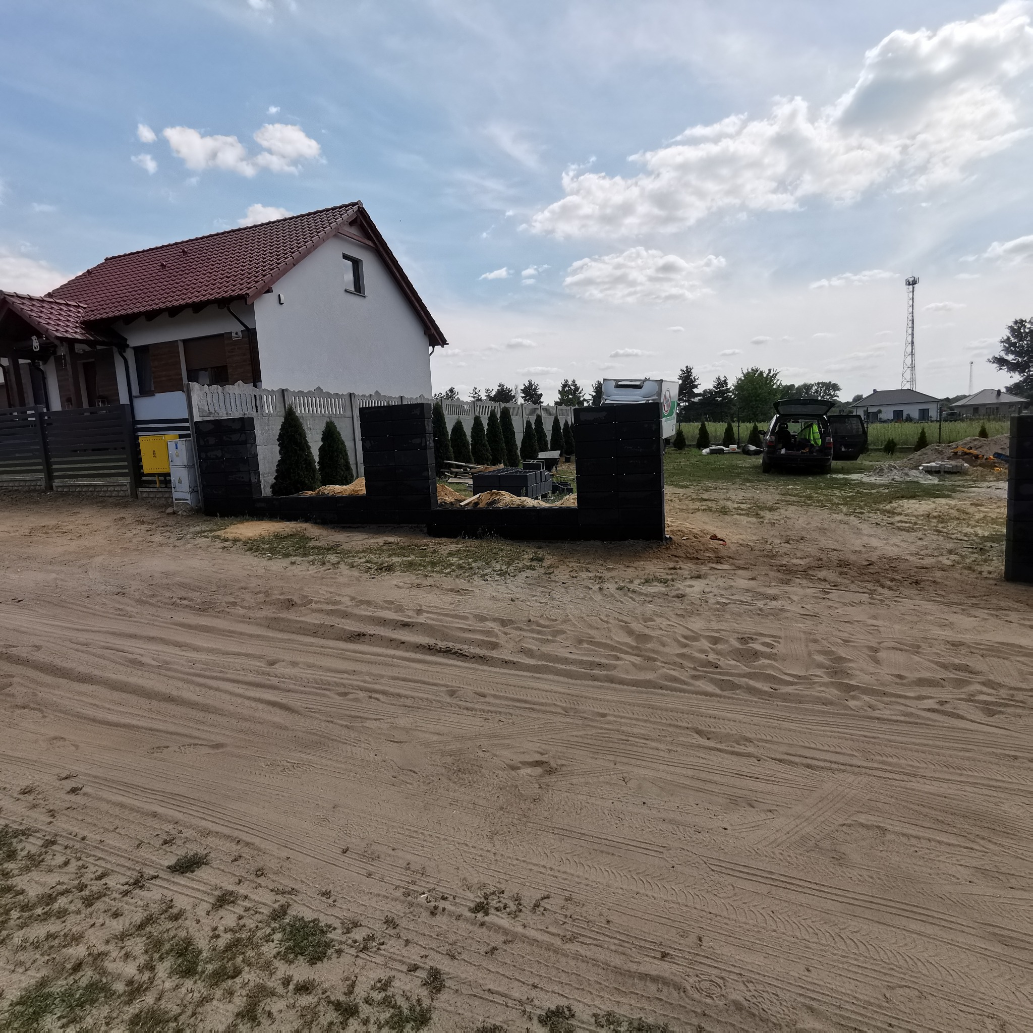 Wznoszenie ogrodzenia betonowego z czarnych bloczków na piaszczystej posesji, widoczny fragment domu i samochód ekipy montażowej w tle.