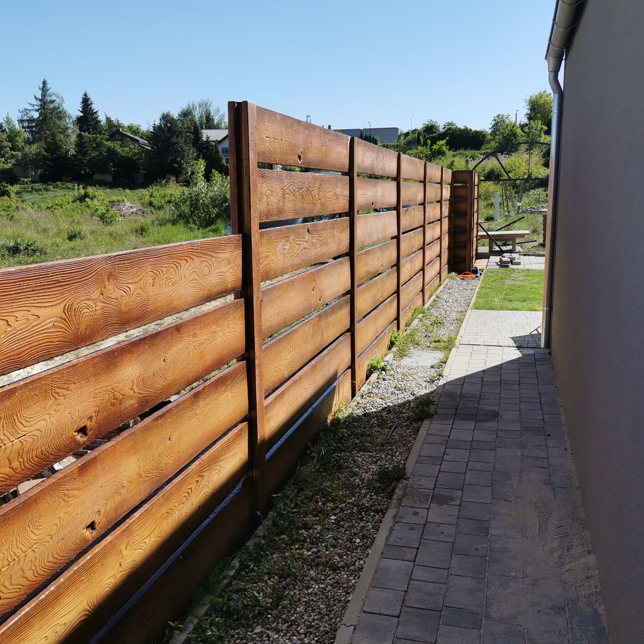 Drewniany płot panelowy z widoczną fakturą słojów, oddzielający posesję od zieleni, z chodnikiem z kostki brukowej wzdłuż ogrodzenia. Słoneczny dzień.