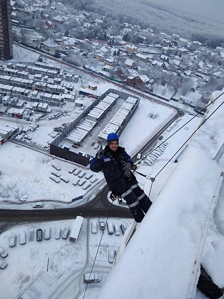 Pracownik na linie, w kasku i kombinezonie, daje znak OK na tle zimowego krajobrazu z budynkami. Praca na wysokości w warunkach zimowych.