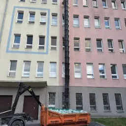 Pomarańczowy kontener wypełniony gruzem podłączony do rury zsypowej przy budynku w Jedlińsku. Widoczny fragment ciężarówki z dźwigiem.