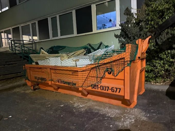 Pomarańczowy kontener z gruzem, zabezpieczony siatką, stoi na zewnątrz budynku. Widoczny numer telefonu i napis 'Wynajem Kontenerów'.