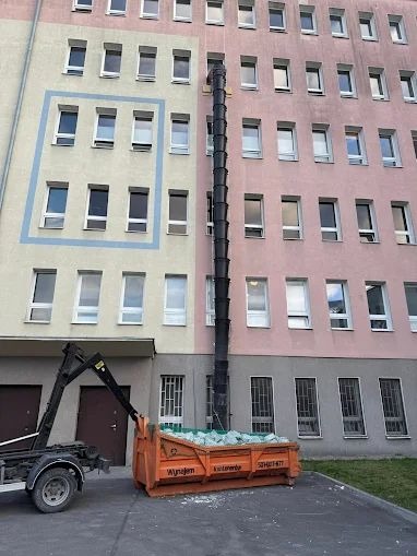 Pomarańczowy kontener wypełniony gruzem podłączony do rury zsypowej przy budynku w Jedlińsku. Widoczny fragment ciężarówki z dźwigiem.