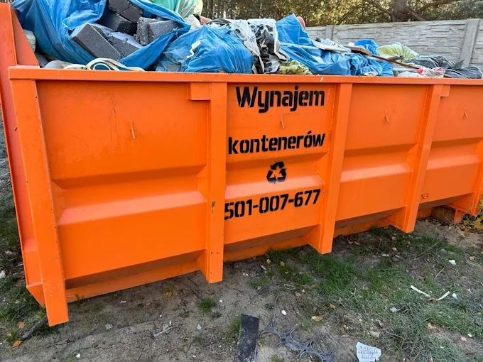 Pomarańczowy kontener na gruz wypełniony odpadami budowlanymi i folią, z napisem 'Wynajem kontenerów' i numerem telefonu na tle zielonej trawy.