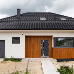 MI-TA group Sp.z.o.o. - Biały dom jednorodzinny z czarnym dachem i drewnianą elewacją, widok z przodu w pochmurny dzień. Nowoczesna architektura z garażem i oknami dachowymi.