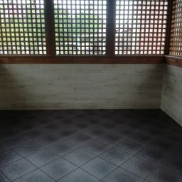 MI-TA group Sp.z.o.o. - Wykończony balkon z brązową kratką, beżowymi płytkami ściennymi i ciemną podłogą z płytek. Widok na zewnątrz przez kratkę.