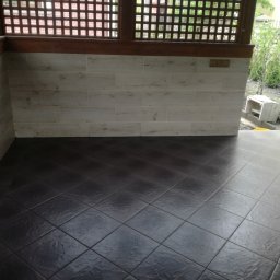 MI-TA group Sp.z.o.o. - Wykończony taras z ciemną, kwadratową podłogą z płytek i jasnymi, poziomymi płytkami na ścianie, widok na ogród przez drewnianą kratkę.