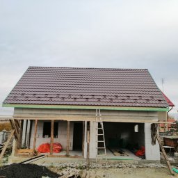 MI-TA group Sp.z.o.o. - Dom w trakcie budowy: brązowy dach z blachodachówki, białe ściany z bloczków. Widoczne rusztowania i drabina. Prace dekarskie w toku.