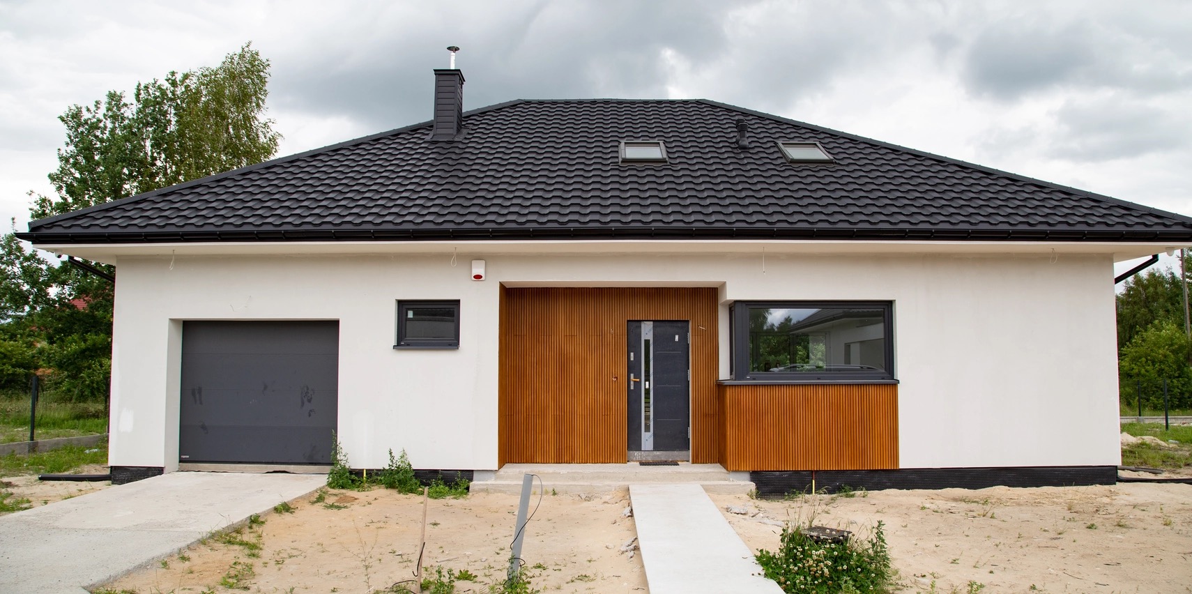 Biały dom jednorodzinny z czarnym dachem i drewnianą elewacją, widok z przodu w pochmurny dzień. Nowoczesna architektura z garażem i oknami dachowymi.