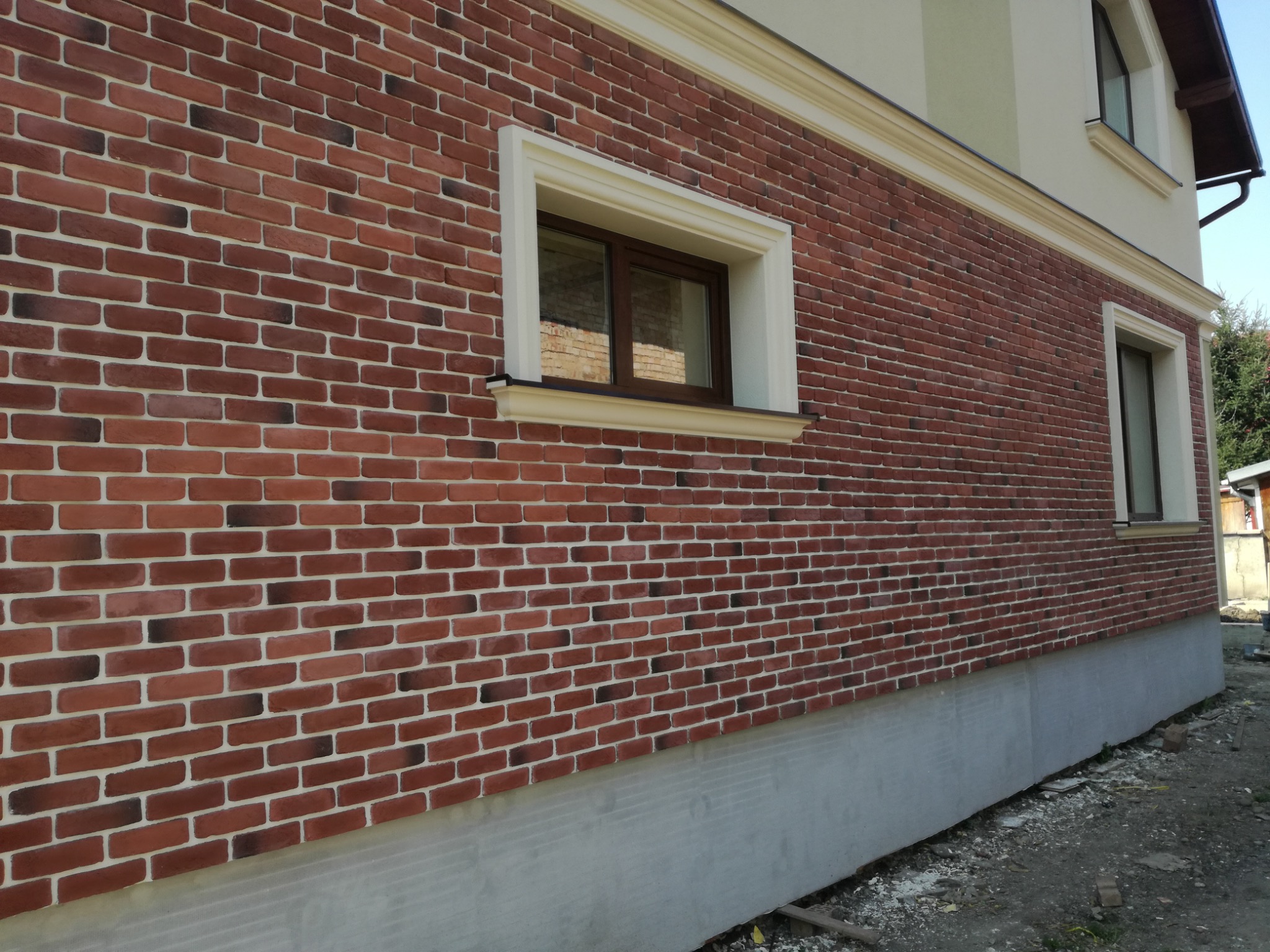 Elewacja budynku wykończona czerwoną cegłą klinkierową z jasną fugą, okna z kremowymi obramowaniami i dekoracyjny gzyms. Widoczne fundamenty.