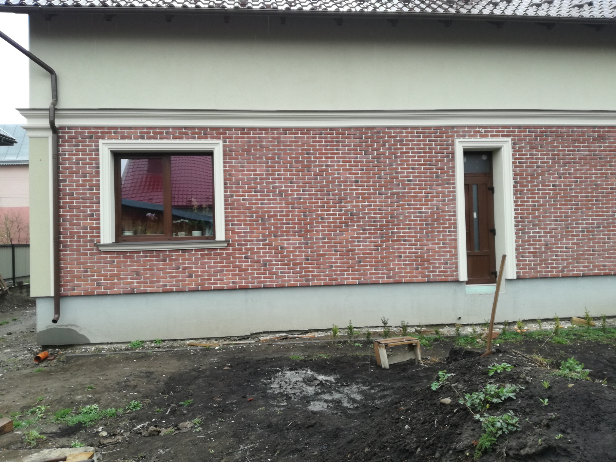 Elewacja budynku z cegły klinkierowej, okno z drewnianą ramą i brązowe drzwi. Widoczny fragment rynny i fundamentu. Na pierwszym planie ziemia.