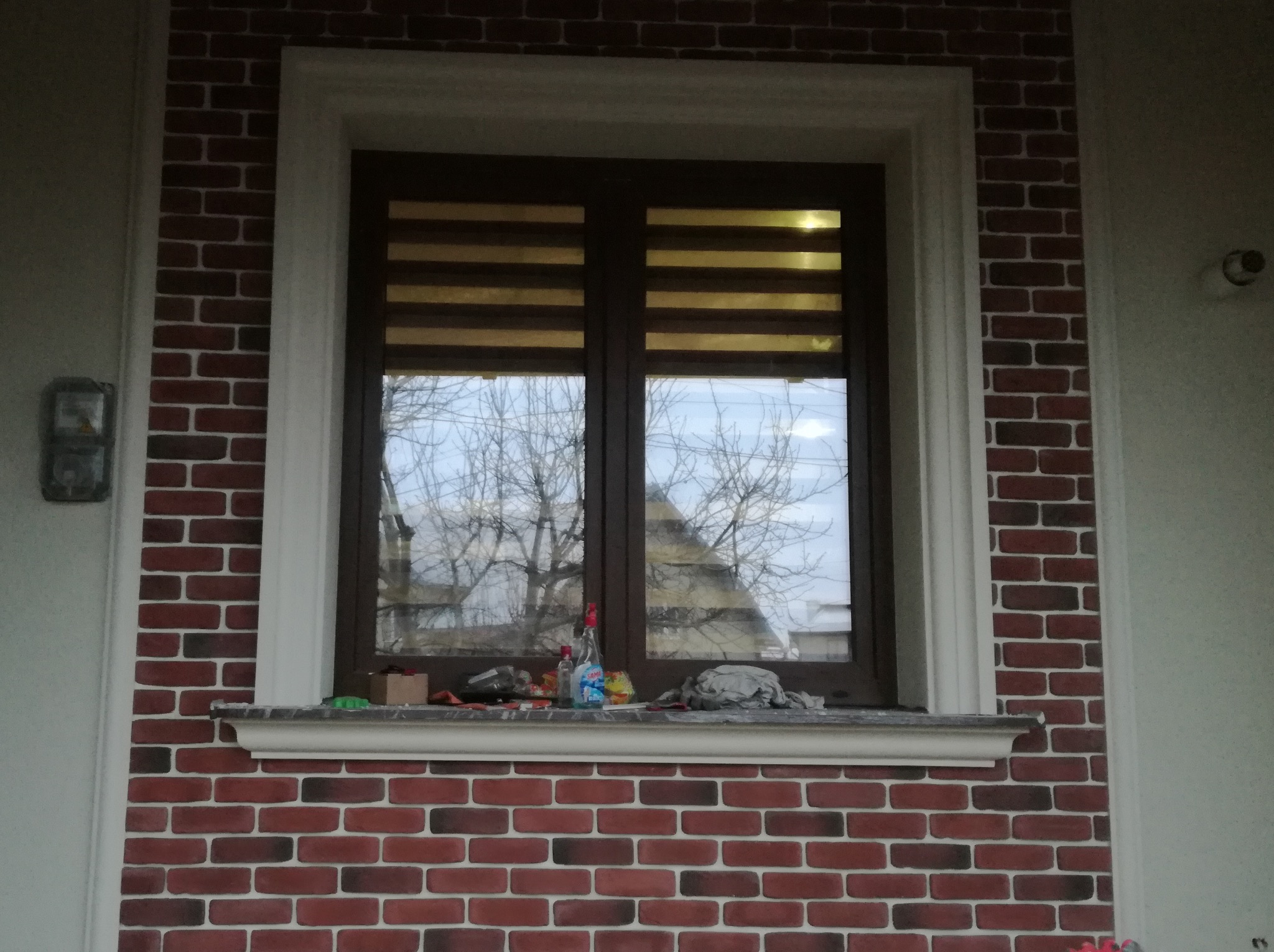 Okna z roletami dzień-noc w elewacji imitującej czerwoną cegłę z białymi fugami, wykończone białym obramowaniem. Na parapecie butelki i inne przedmioty.