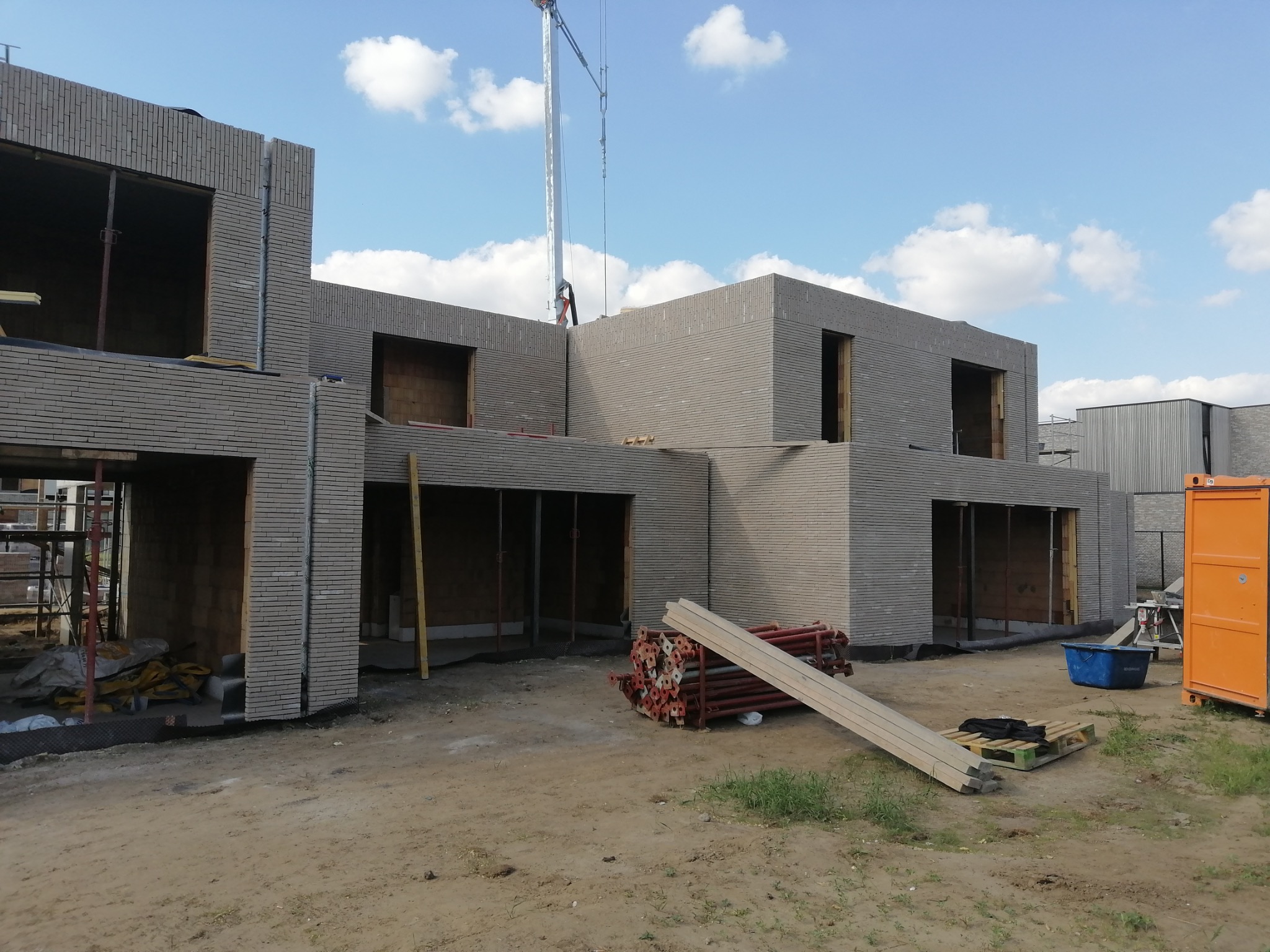 Surowy stan budynku z szarej cegły, widoczny żuraw budowlany i materiały budowlane. Budowa w trakcie, jasne niebo z chmurami w tle.