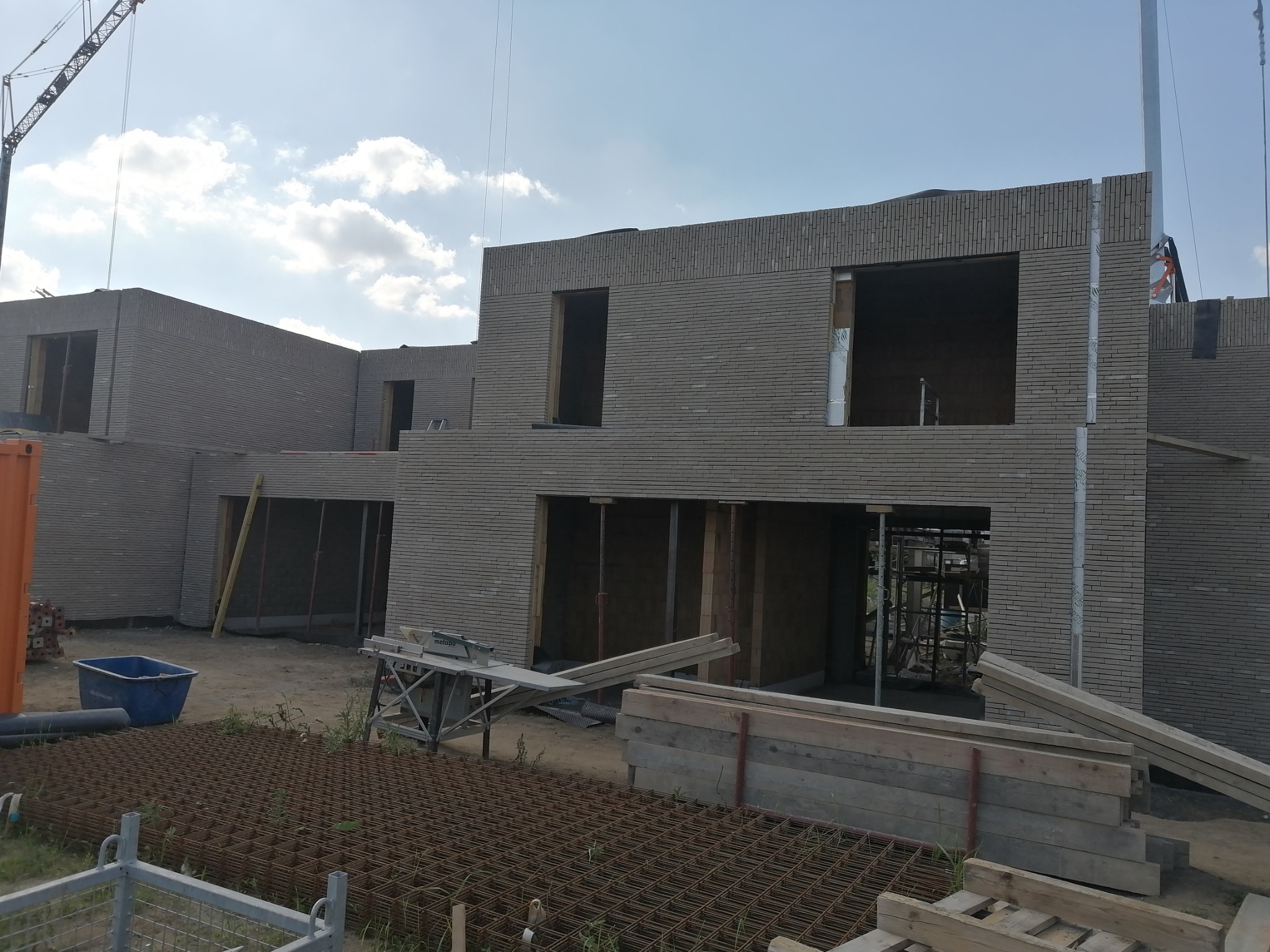 Wznoszenie surowego budynku z szarej cegły. Widoczne otwory okienne i drzwiowe, rusztowania wewnątrz, materiały budowlane na pierwszym planie. Na horyzoncie dźwig.