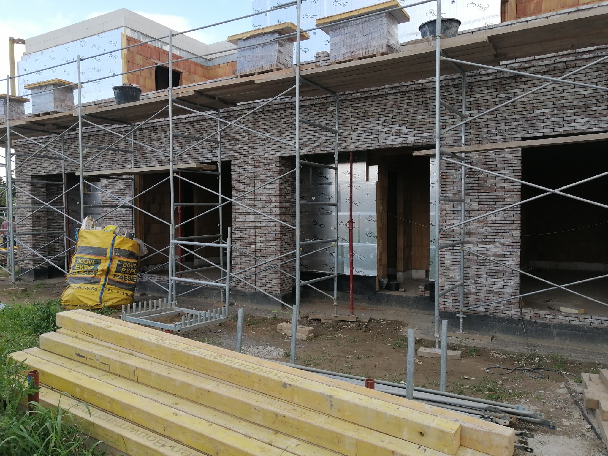 Budowa domu z cegły klinkierowej w trakcie realizacji. Widoczne rusztowania, materiały budowlane i fragmenty ocieplenia. Cegły na paletach na rusztowaniu.