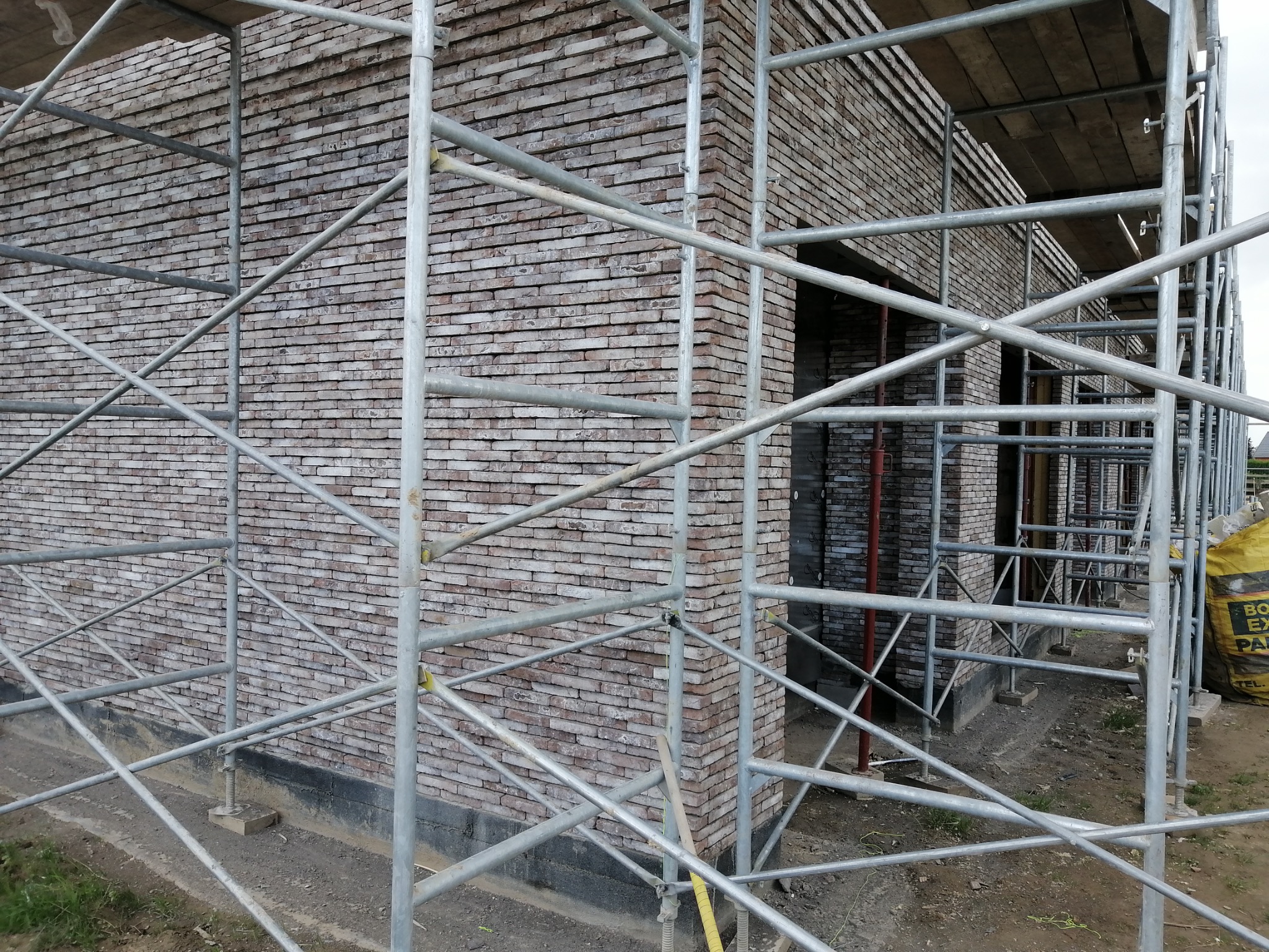 Murowana elewacja budynku w trakcie budowy, widoczna przez rusztowanie. Jasna cegła, poziomy układ. Fragment worka z zaprawą w tle.