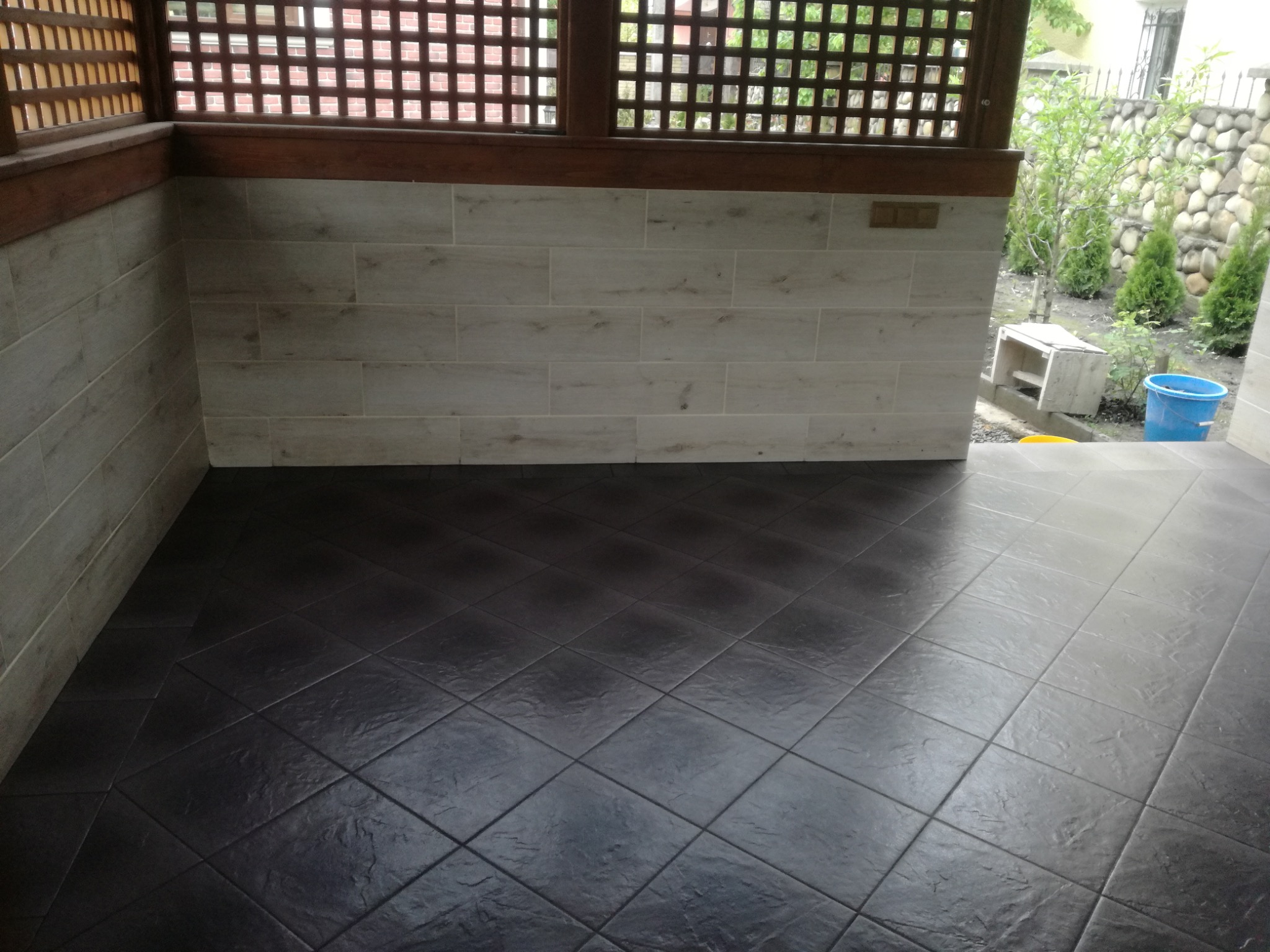 Wykończony taras z ciemną, kwadratową podłogą z płytek i jasnymi, poziomymi płytkami na ścianie, widok na ogród przez drewnianą kratkę.