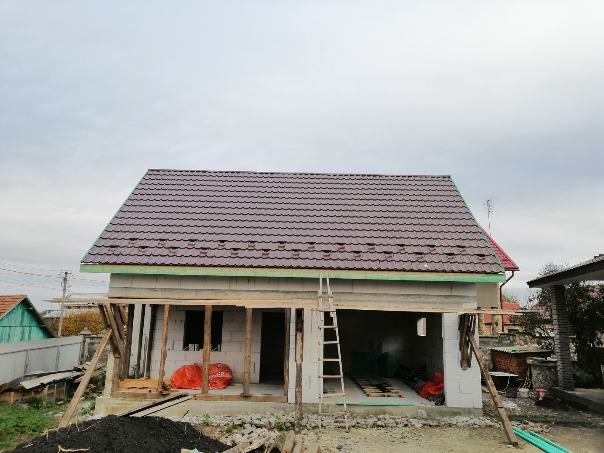 Dom w trakcie budowy: brązowy dach z blachodachówki, białe ściany z bloczków. Widoczne rusztowania i drabina. Prace dekarskie w toku.