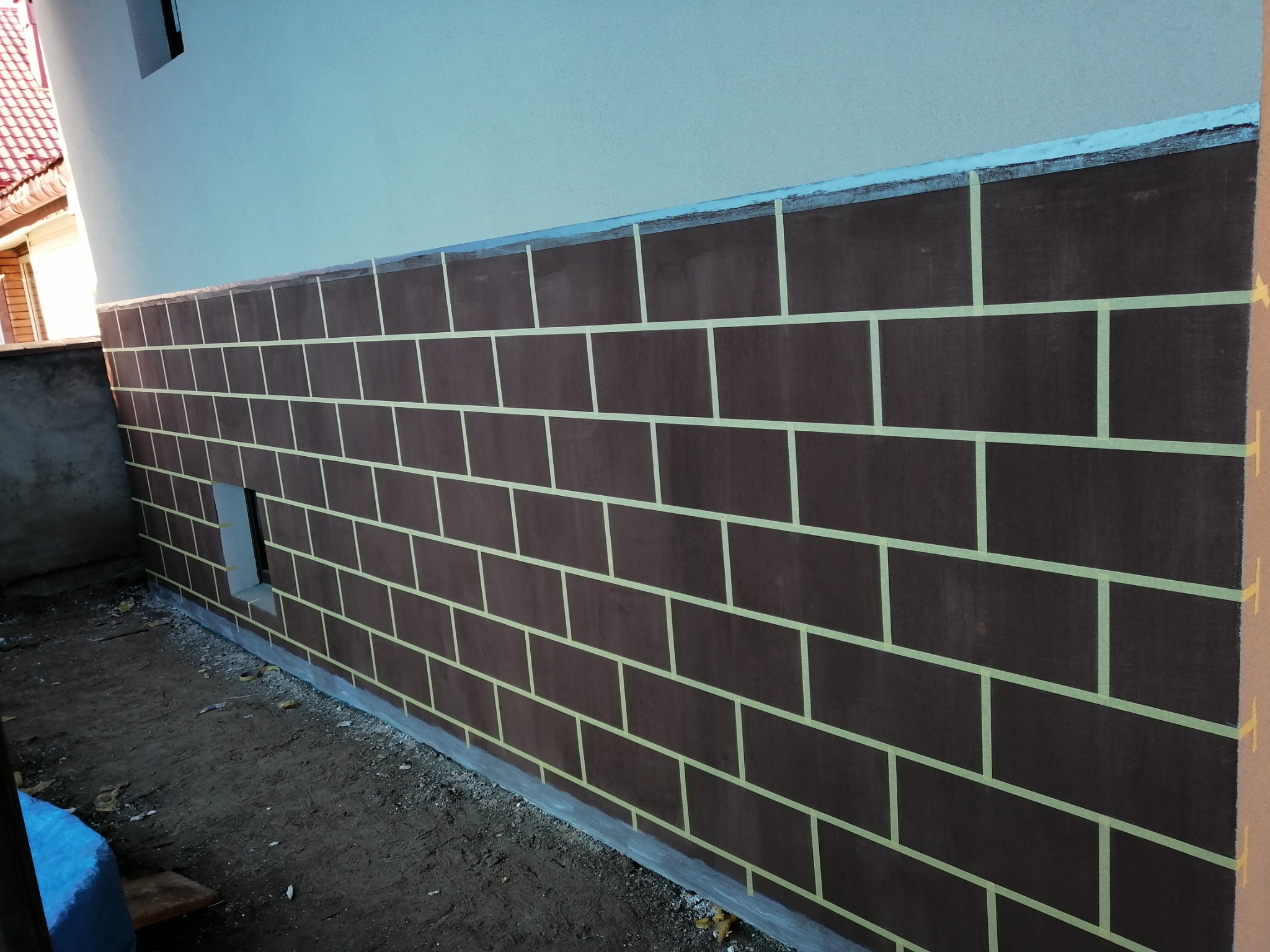 Malowanie elewacji imitujące cegłę, z użyciem taśmy malarskiej. Brązowa ściana z regularnym wzorem cegieł, częściowo widoczny budynek w tle.