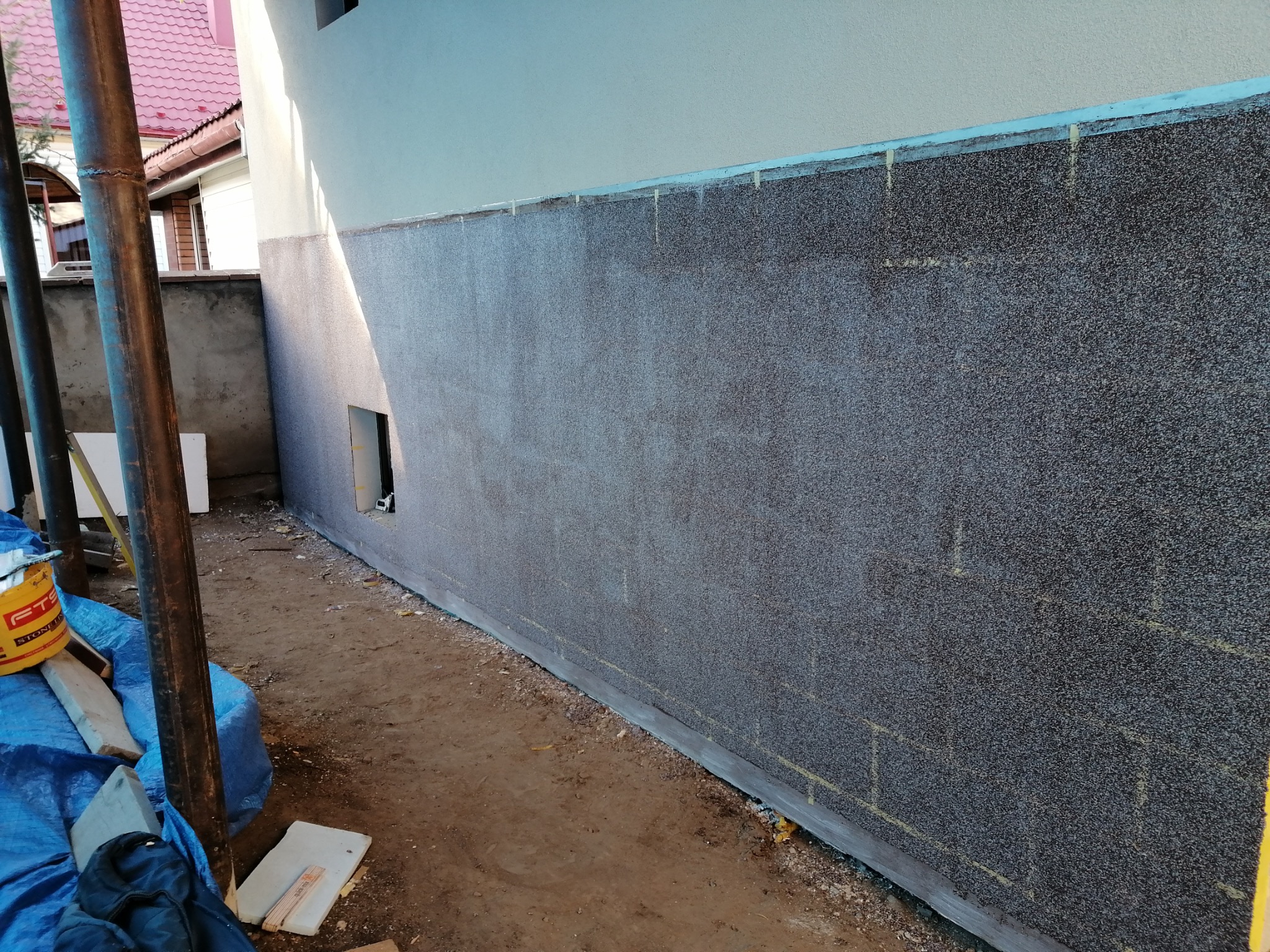 Elewacja domu w trakcie wykończenia, z nałożoną warstwą dekoracyjną w kolorze szaro-brązowym. Widoczne rusztowania i materiały budowlane.