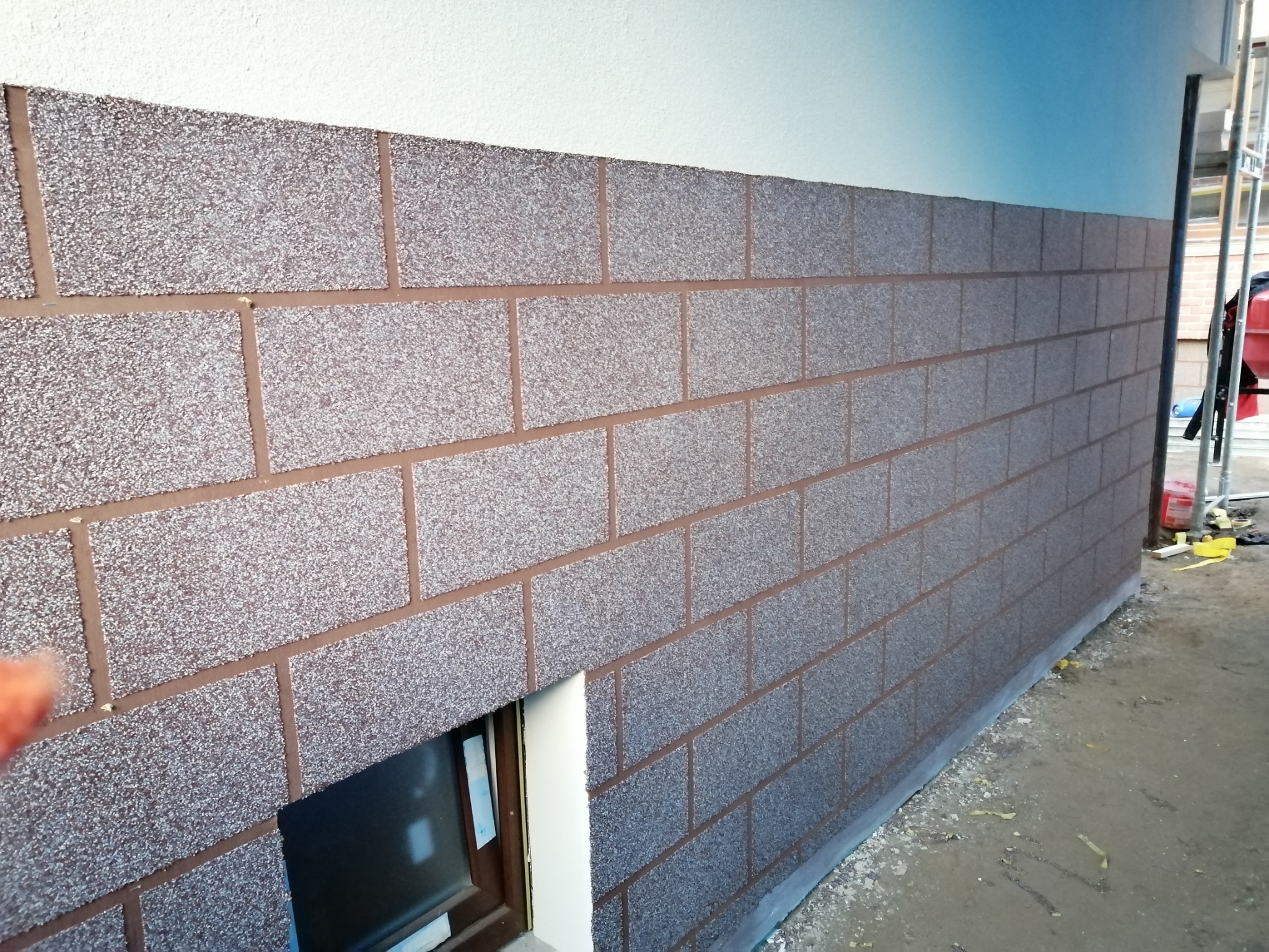 Elewacja z dekoracyjnym tynkiem imitującym cegłę, fragment okna piwnicznego, widoczne rusztowanie w tle. Teksturowana ściana w odcieniach szarości i brązu.