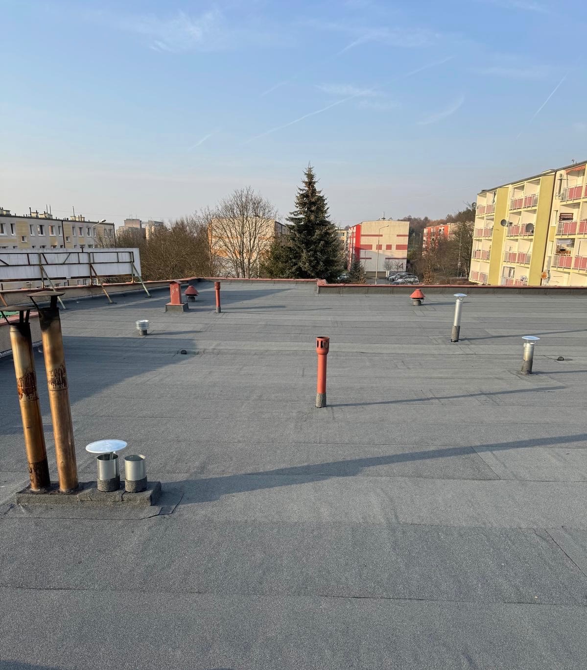 Szary, płaski dach pokryty papą termozgrzewalną z kominami wentylacyjnymi i metalowymi zakończeniami. W tle budynki mieszkalne i drzewa w słoneczny dzień.