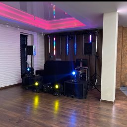 Mateusz Zabiegły - Sala z parkietem, przygotowana na imprezę. Widoczne nagłośnienie, oświetlenie sceniczne, reflektory i dekoracje świetlne na suficie. Gotowość do wydarzenia.