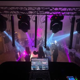 Mateusz Zabiegły - DJ na imprezie z oświetleniem: laptop z mikserem, reflektory, kolumny, tańczący ludzie. Fioletowe i niebieskie światła tworzą klimat. Widok z perspektywy DJ-a.