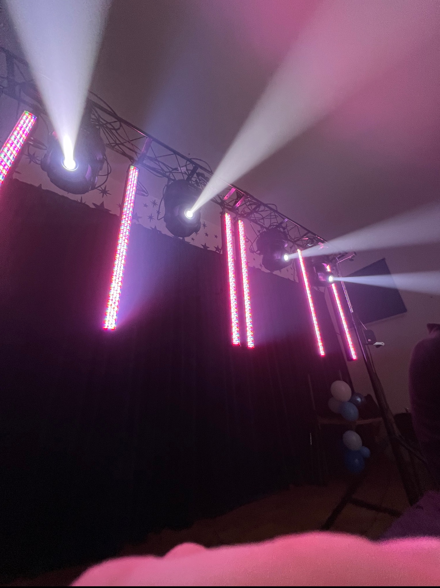 Scena oświetlona reflektorami i pionowymi lampami LED w kolorze różowym i białym, z elementami dekoracji i balonami w tle. Oświetlenie sceniczne.