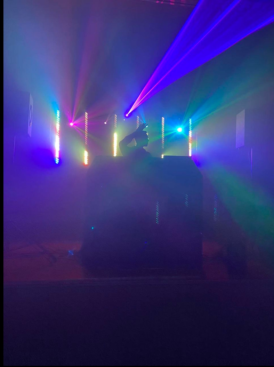 Sylwetka DJ-a w dymie, podświetlona kolorowymi laserami i światłami LED. Intensywne, barwne oświetlenie sceniczne tworzy dynamiczną atmosferę imprezy.