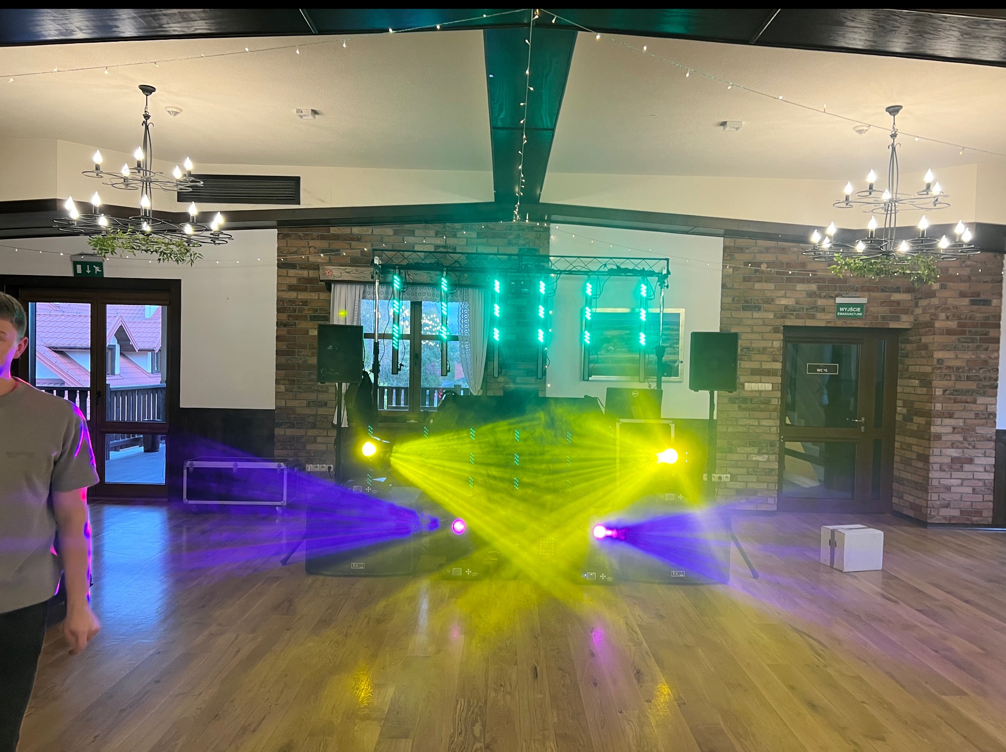 Sala z parkietem, oświetleniem estradowym i nagłośnieniem. Widoczne kolumny głośnikowe, reflektory generujące żółte i fioletowe światło, konstrukcja oświetleniowa.