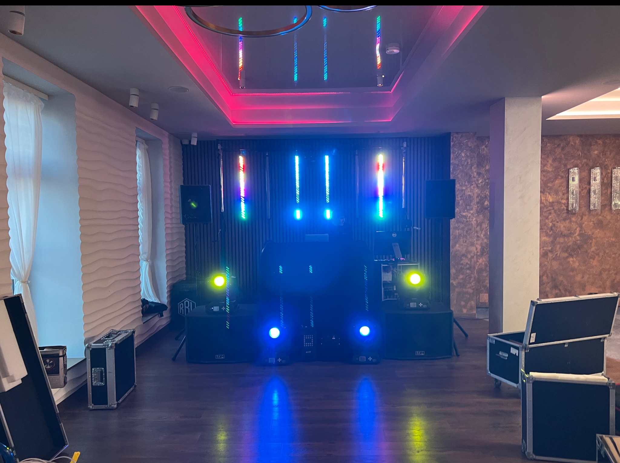 Sala z nagłośnieniem i oświetleniem gotowa na imprezę. Widoczne kolumny, reflektory i skrzynie transportowe. Na suficie dekoracyjne oświetlenie LED.