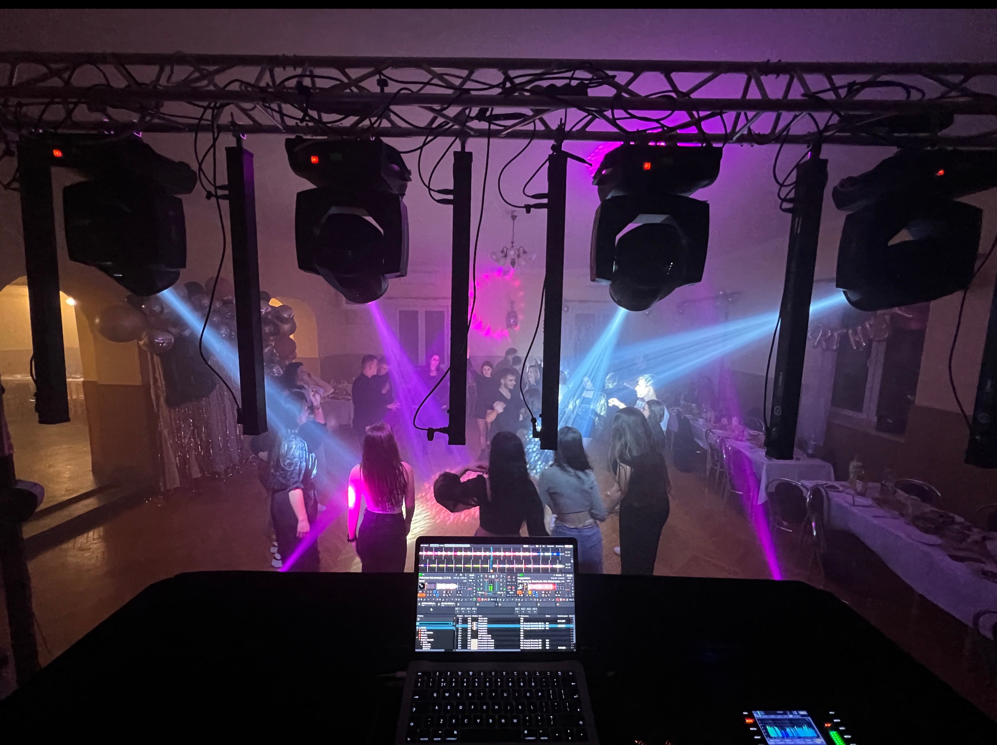 DJ na imprezie z oświetleniem: laptop z mikserem, reflektory, kolumny, tańczący ludzie. Fioletowe i niebieskie światła tworzą klimat. Widok z perspektywy DJ-a.