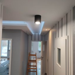 Adam Kaczmarek - Pomalowany korytarz w odcieniach szarości i bieli, z drewnianą drabiną, widoczne oświetlenie sufitowe i wzorzysta tapeta. W tle otwarte drzwi do kolejnych pomieszczeń.