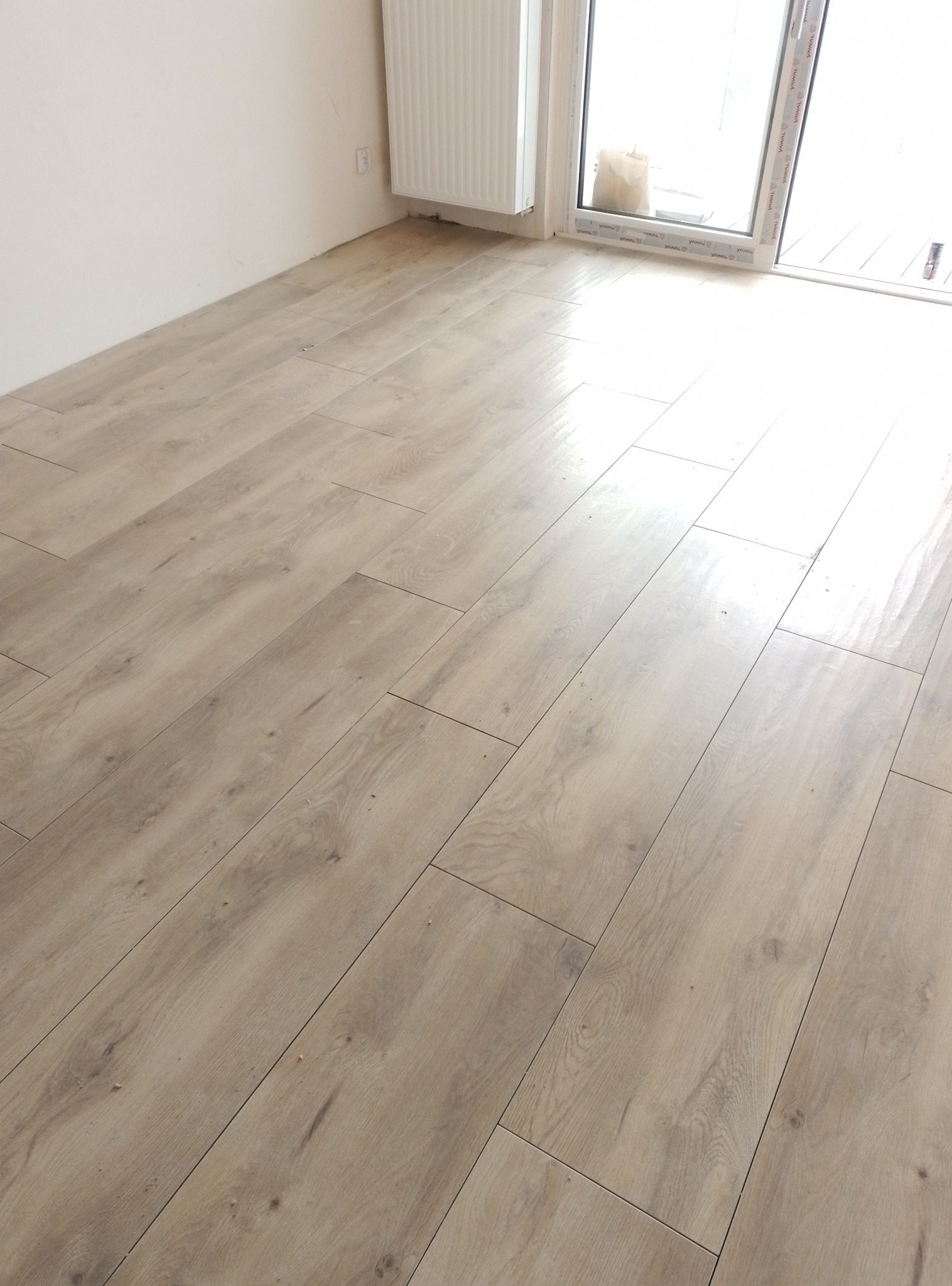 Ułożona podłoga z paneli imitujących drewno, widok z perspektywy narożnika pokoju z grzejnikiem i oknem balkonowym w tle. Jasne wnętrze w stanie deweloperskim.