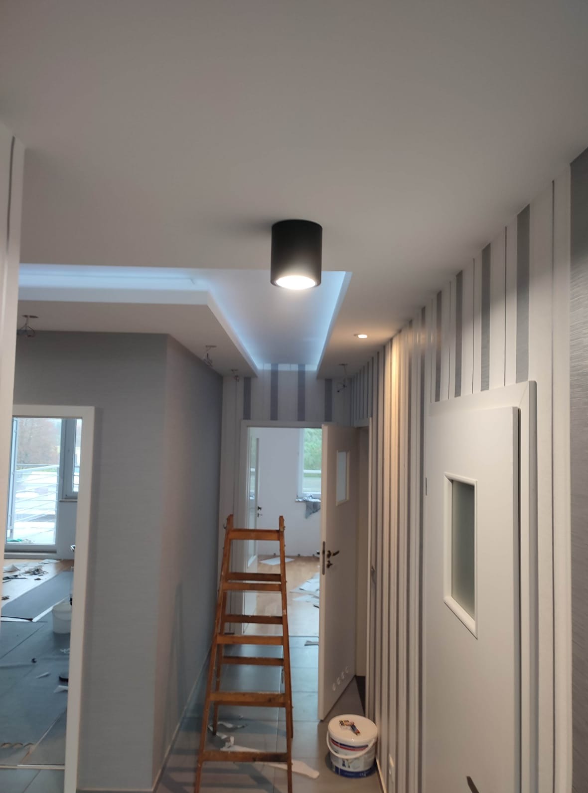 Pomalowany korytarz w odcieniach szarości i bieli, z drewnianą drabiną, widoczne oświetlenie sufitowe i wzorzysta tapeta. W tle otwarte drzwi do kolejnych pomieszczeń.