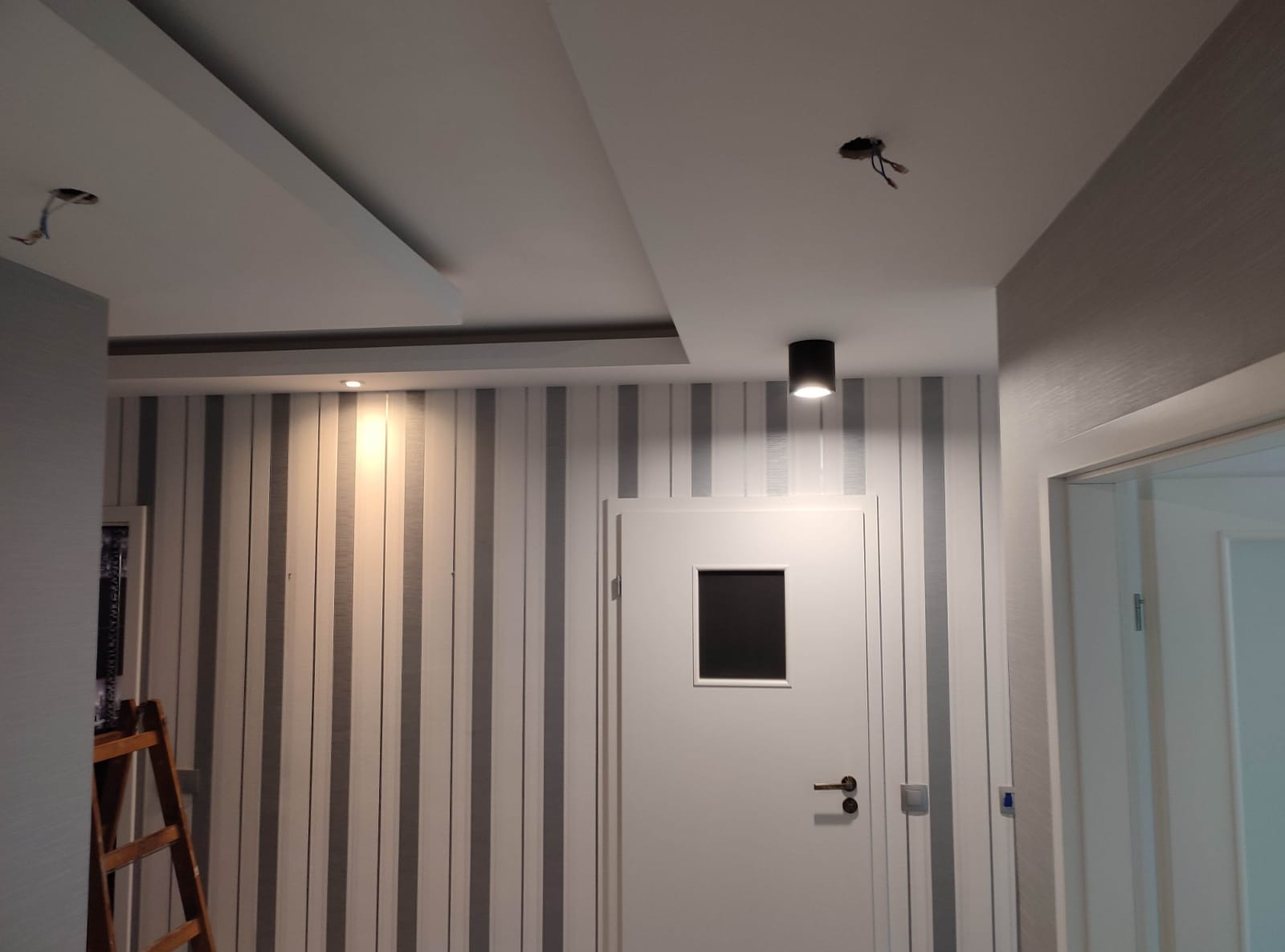 Pionowe pasy na ścianie, białe drzwi z czarnym okienkiem, czarna lampa sufitowa. Widoczne fragmenty sufitu z elementami instalacji elektrycznej.