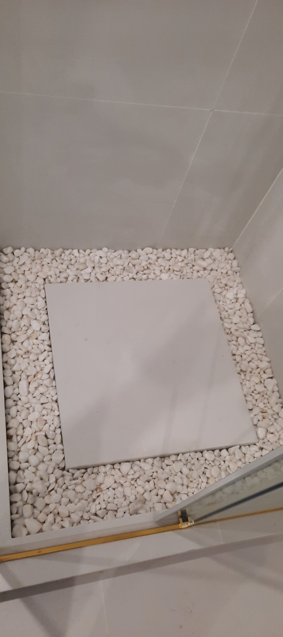 Wykończenie brodzika z białych płytek i otoczaków, widoczne złote detale kabiny prysznicowej. Minimalistyczny design w łazience.