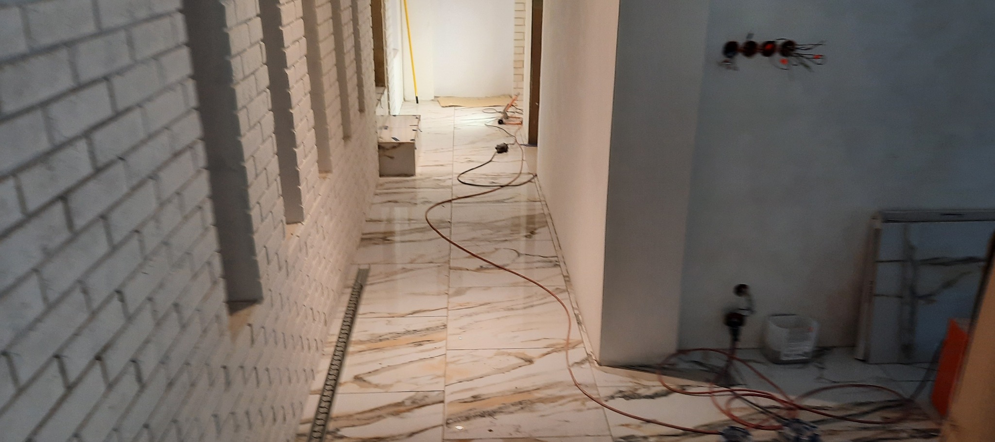 Korytarz w trakcie prac wykończeniowych: położona marmurowa podłoga, ściana z białej cegły dekoracyjnej, widoczne kable i narzędzia.