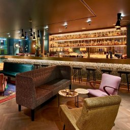 Kamil Konkol Fotograf Nieruchomości Wrocław, wnętrz, domów, mieszkań, Wrocław - Eklektyczny bar z mozaikowym blatem, oświetlonymi półkami pełnymi butelek, pluszowymi sofami i fotelami w odcieniach różu i złota na parkiecie ułożonym we wzór jodełki.