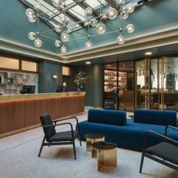 Kamil Konkol Fotograf Nieruchomości Wrocław, wnętrz, domów, mieszkań, Wrocław - Recepcja hotelowa z drewnianą ladą, niebieską sofą i szklanym sufitem z designerską lampą. W tle widoczny bar za szklanymi drzwiami.